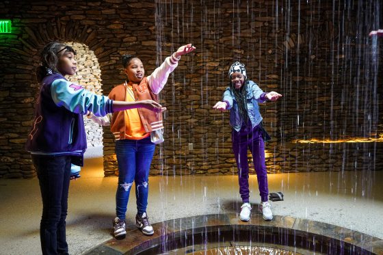 Three people standing around a fountain putting their hands in it.